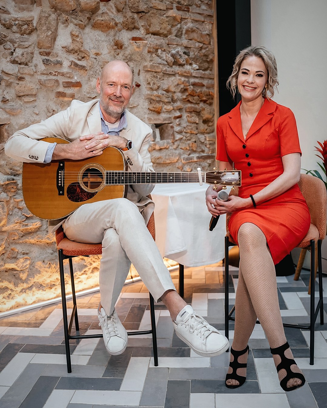 Geir Løken with his guitar and Eva Iselinn Erichsen with a microphone, seated in front of an old stone wall